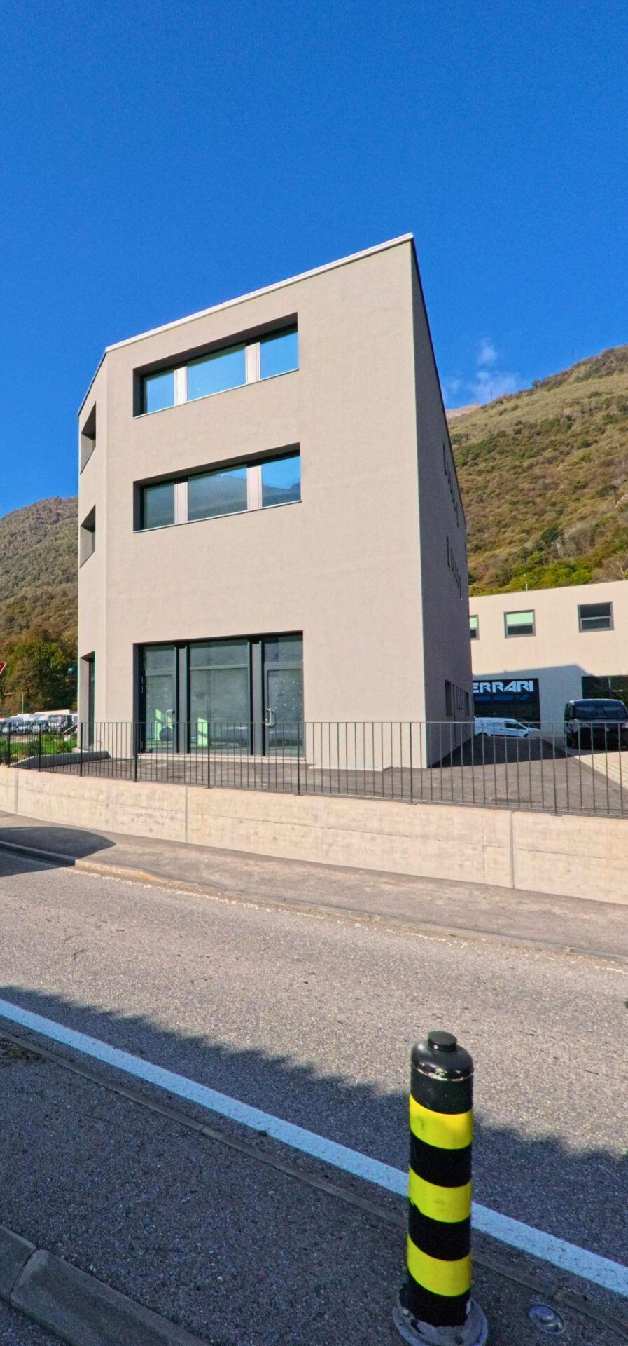 Vor einem bewaldeten Hang steht ein modernes, kubisches Gebäude entlang der Strasse in Mezzovico-Vira (6805, Kanton Tessin) unter klarem blauem Himmel.