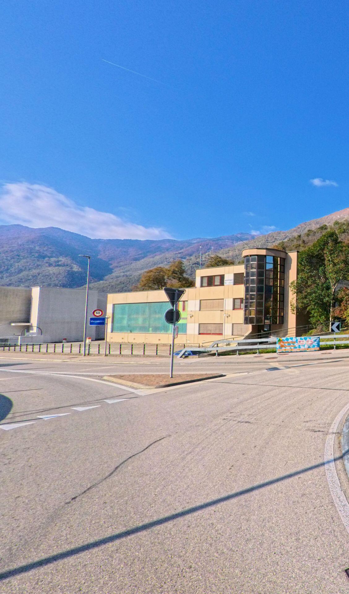 An einem Verkehrsknotenpunkt steht ein markantes Gebäude am Rand von Mezzovico-Vira (6805, Kanton Tessin), vor dem sich die Strasse öffnet und bewaldete Berghänge den Hintergrund bilden.