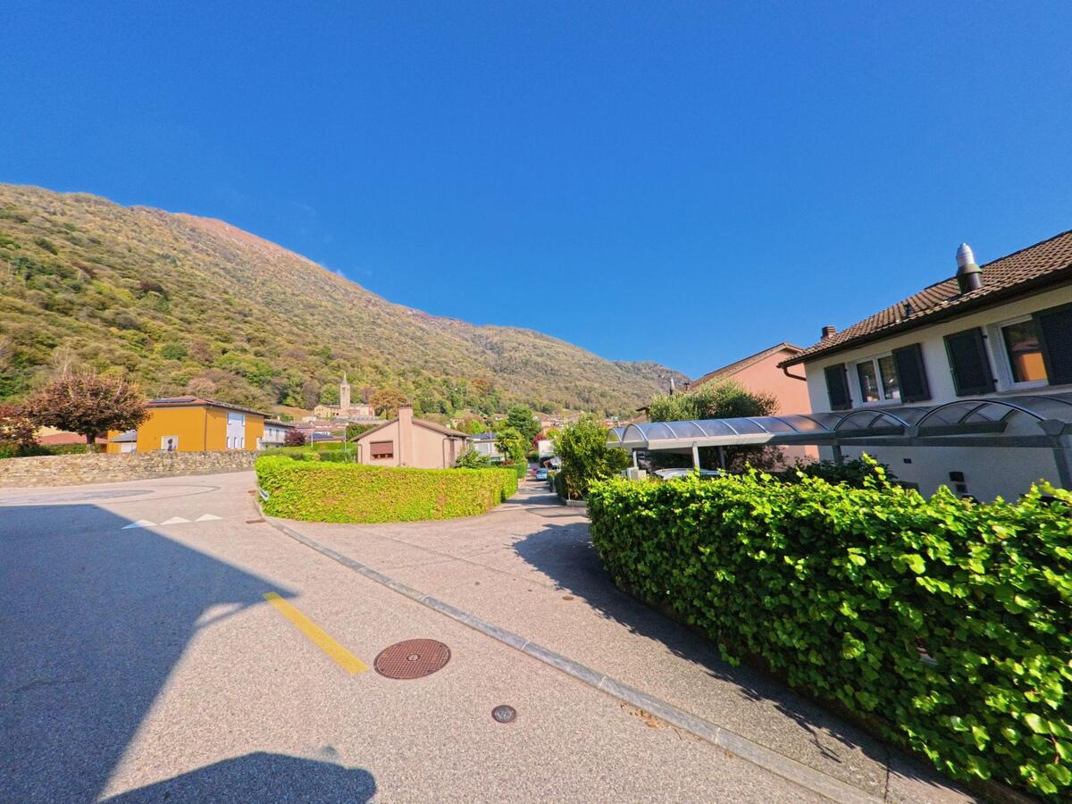 Entlang einer ruhigen Zufahrt mit gepflegten Hecken führt der Blick auf Wohnhäuser in Mezzovico-Vira (6805, Kanton Tessin), während sich dahinter sanft ansteigende Berghänge abzeichnen.