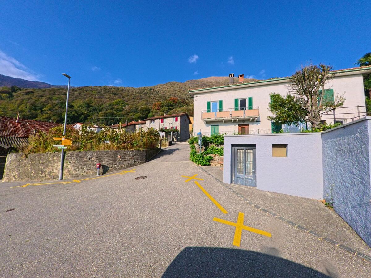An einer ruhigen Einmündung mit markierten Parkflächen verläuft eine schmale Strasse zwischen Wohnhäusern in Mezzovico-Vira (6805, Kanton Tessin), während sich dahinter bewaldete Hänge erheben.