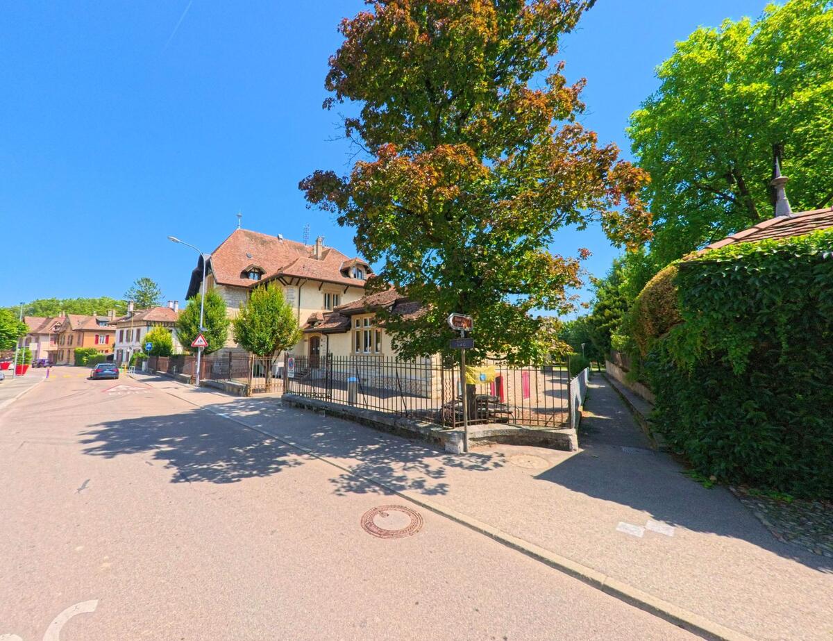 In Milvignes (2012, Kanton Neuenburg) verläuft eine ruhige Strasse mit historischen Wohnhaeusern und grossen Baeumen unter blauem Himmel.