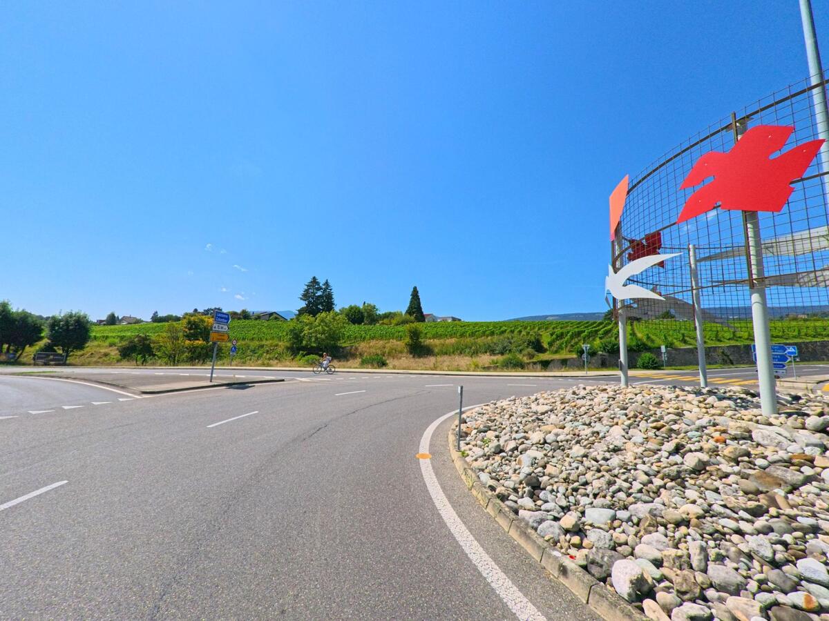 Ein Kreisverkehr in Milvignes (2012, Kanton Neuenburg) ist mit einem Metallgitter und farbigen Formen gestaltet, im Hintergrund liegen Weinreben an einem sonnigen Hang.
