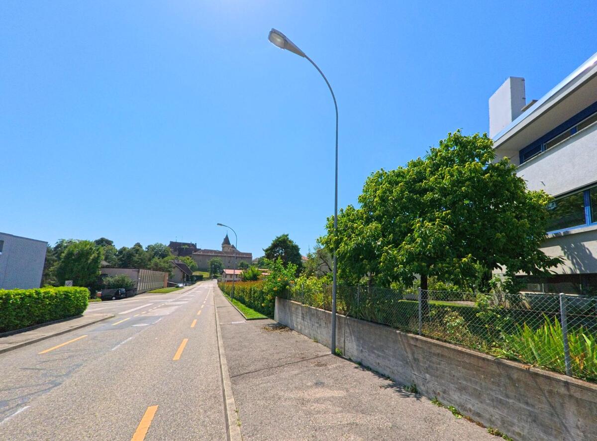 Eine gerade Strasse in Milvignes (2013, Kanton Neuenburg) fuehrt zwischen modernen Wohnhaeusern und Baeumen in Richtung des Kirchturms im Hintergrund.