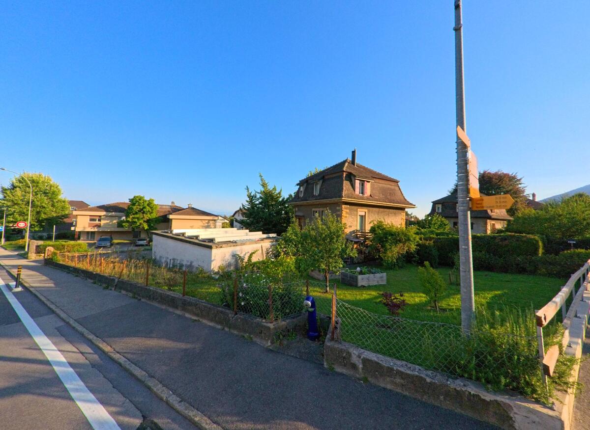 Ein Einfamilienhaus mit Mansardendach und angrenzendem Garten liegt in Milvignes (2014, Kanton Neuenburg) an einer Strasse mit Fussgaengerweg.
