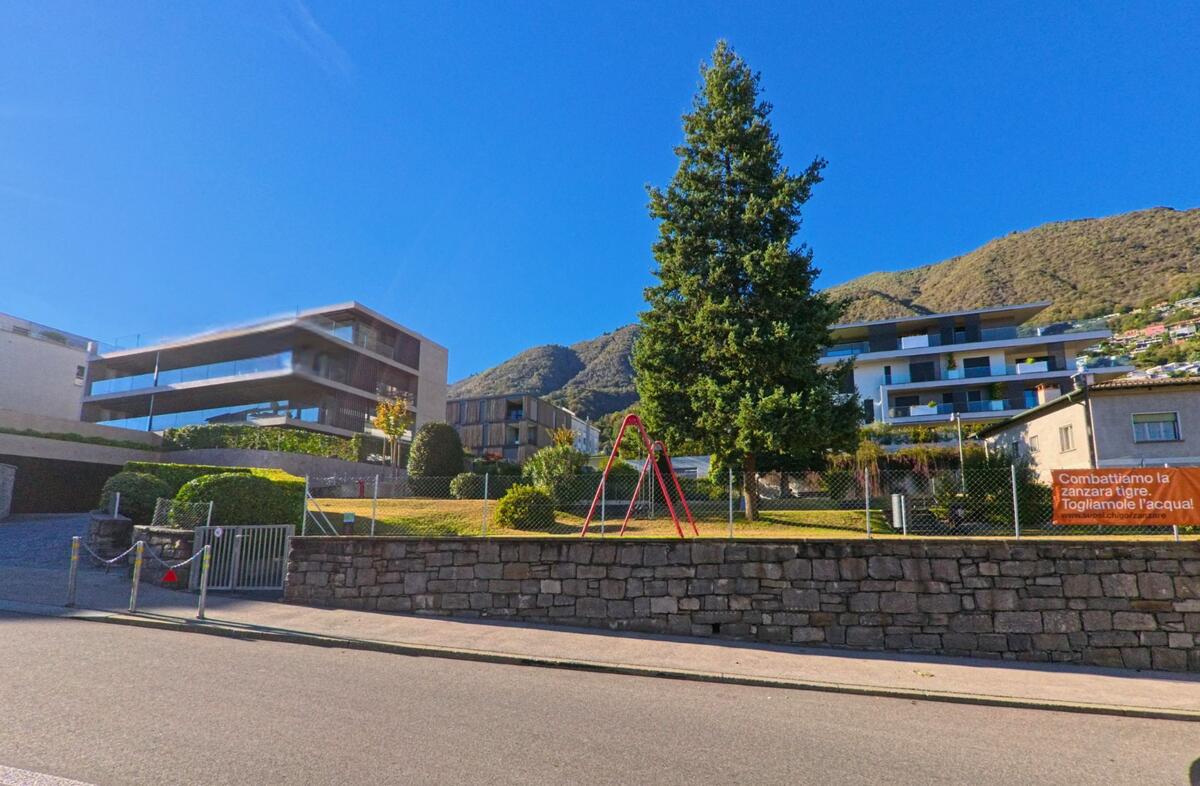Das Gebäude der Casa Comunale in Minusio (6648, Kanton Tessin) steht an einer sonnigen Strasse neben einer Bushaltestelle mit Blick auf die Berge.