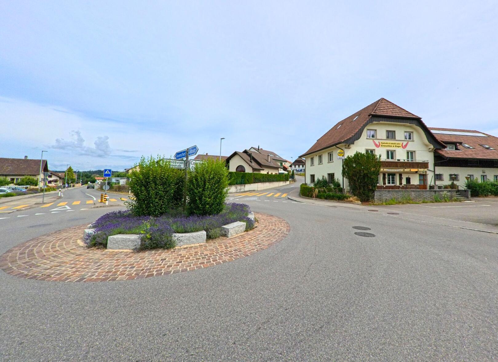 In Misery-Courtion (1721, Kanton Freiburg) zeigt das Bild einen kleinen bepflanzten Kreisverkehr mit Hinweisschildern, flankiert von Wohnhäusern und einem Restaurantgebäude.