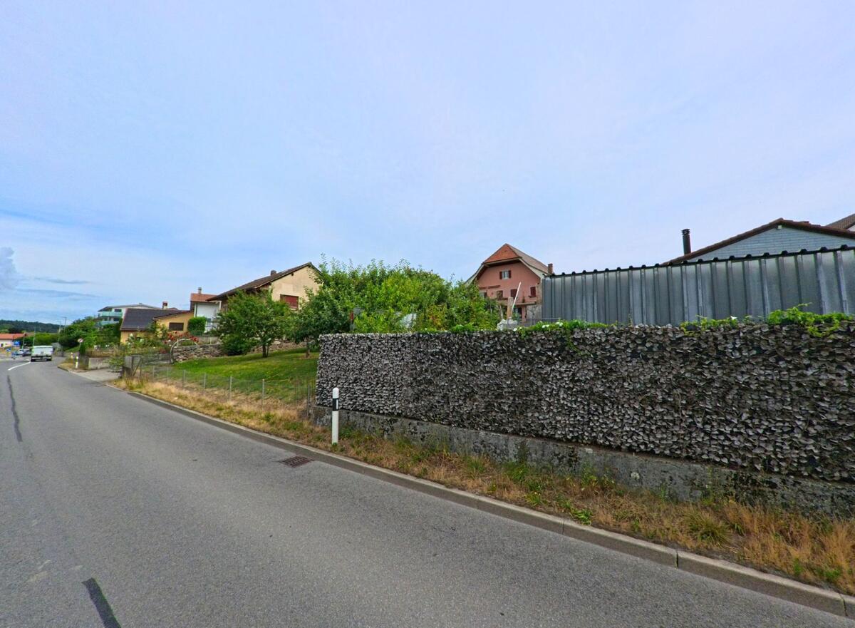 In Misery-Courtion (PLZ 1721, Kanton Freiburg) verläuft eine Nebenstrasse an einer Gabionenmauer vorbei mit Blick auf Gärten und Einfamilienhäuser.