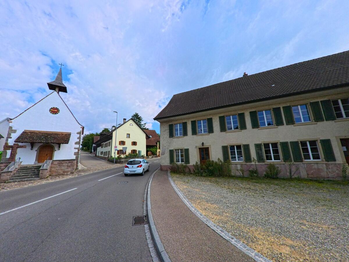 In Möhlin (4313, Kanton Aargau) steht eine kleine weisse Kirche mit Holztor und Turm neben traditionellen Wohnhäusern an einer leicht ansteigenden Strasse.