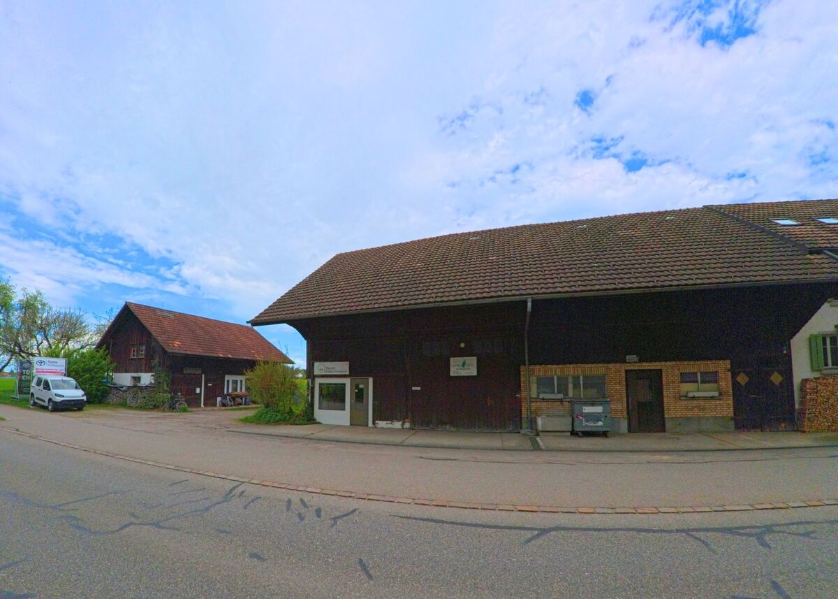 In Mönchaltorf, PLZ 8617 im Kanton Zürich, stehen zwei grosse Bauernhäuser mit dunkler Holzfassade an einer ruhigen Strasse, eines davon mit einem kleinen Hofladen im Eingangsbereich.