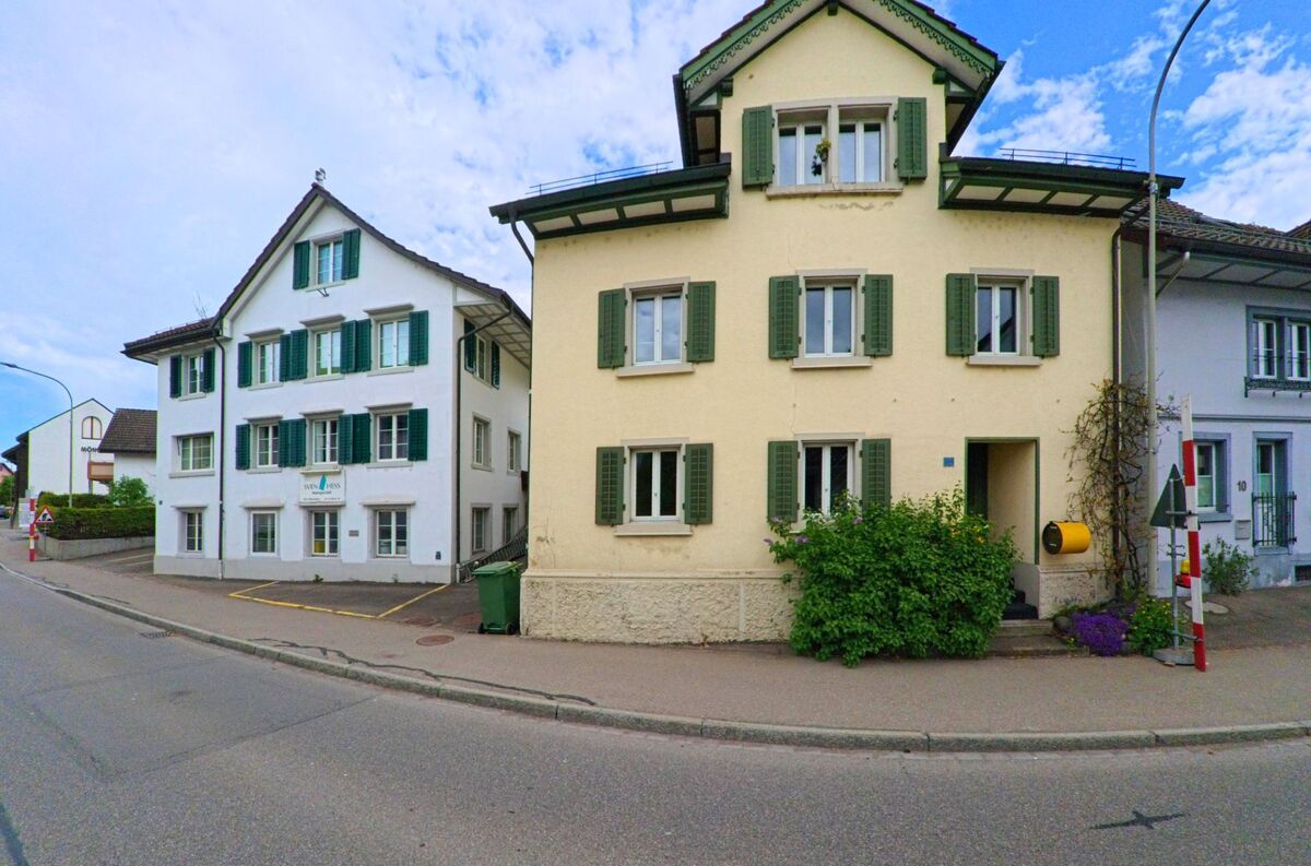 Zwei gepflegte Mehrfamilienhäuser mit grünen Fensterläden stehen im Zentrum von Mönchaltorf, PLZ 8617 im Kanton Zürich, nahe befahrener Strassen und in direkter Nachbarschaft zum Gemeindehaus.
