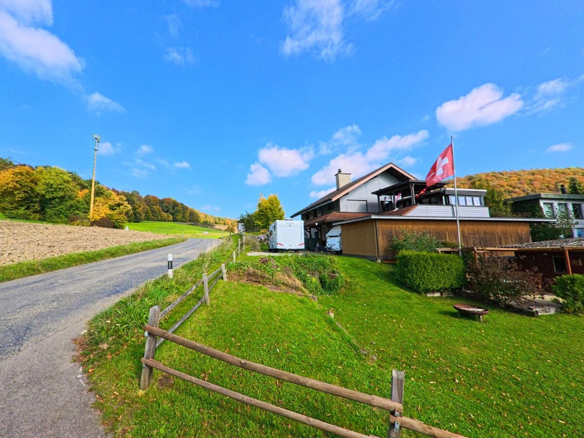 Am Ortsrand von Mönthal (5237, Kanton Aargau) steht ein Wohnhaus mit Schweizer Fahne und Blick auf Felder und herbstlich gefärbte Wälder.