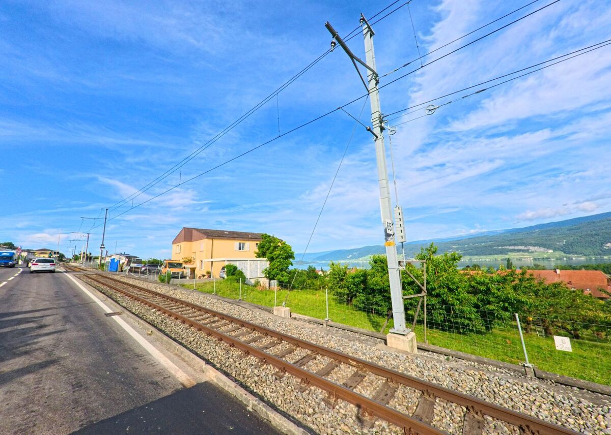 In Mörigen (2572, Kanton Bern) verläuft die Bahnlinie parallel zur Strasse, vorbei an einem gelben Wohnhaus und grüner Vegetation mit Blick auf die Hügel des Juras.