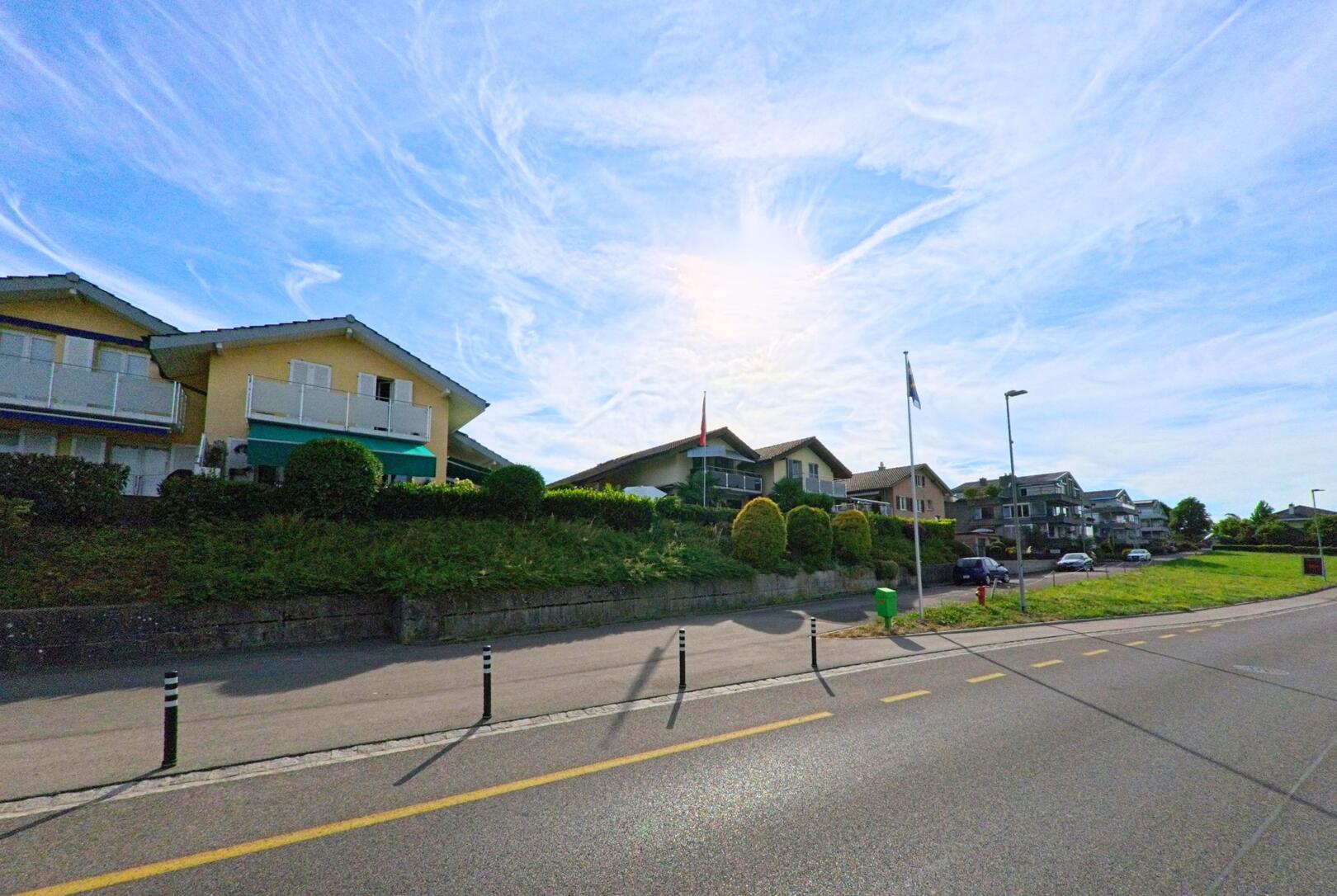 In Mörigen (2572, Kanton Bern) reihen sich Einfamilienhäuser mit gepflegten Vorgärten entlang der leicht ansteigenden Hauptstrasse unter einem weitläufigen Himmel.