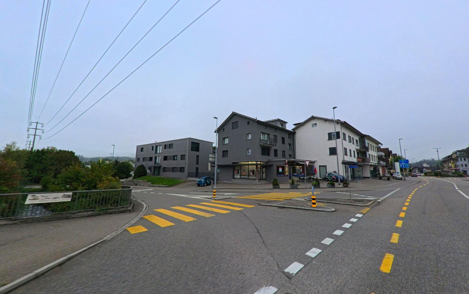 In Möriken-Wildegg (5103, Kanton Aargau) zeigt sich eine Kreuzung mit modernen Wohnhäusern und Geschäften entlang der Hauptstrasse an einem trüben Tag.