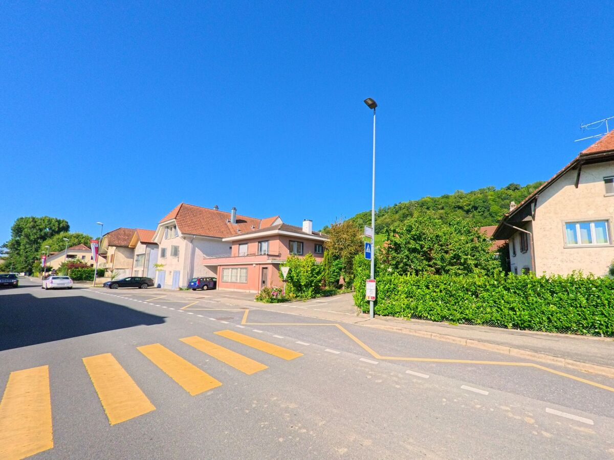 In Mont-Vully (1786, Kanton Freiburg) führt ein Zebrastreifen zu einer Strasse mit Wohnhäusern, hinter denen sich ein bewaldeter Hügel erhebt.