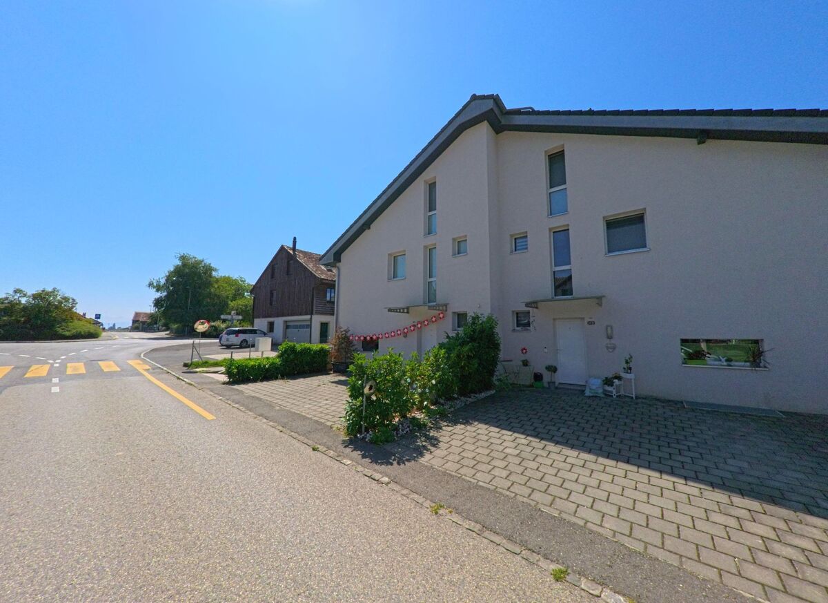 Ein modernes Wohnhaus mit gepflastertem Vorplatz steht in Montanaire (1062, Kanton Waadt) an einer Strassenkreuzung mit Fussgaengerstreifen.