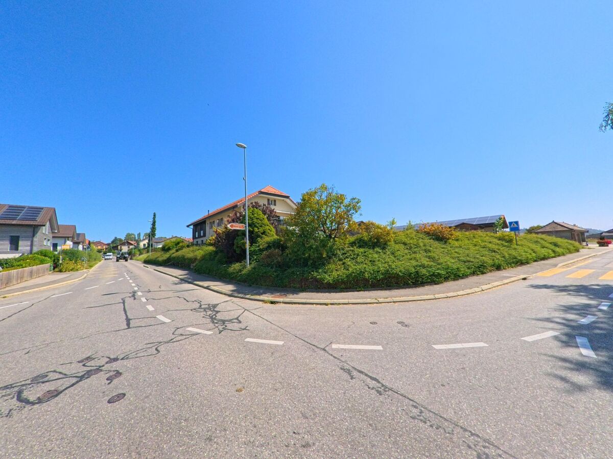 Eine breite Strasse mit Fussgaengerstreifen fuehrt in Montanaire (1062, Kanton Waadt) an Wohnhaeusern mit Gaerten und Solardach vorbei.