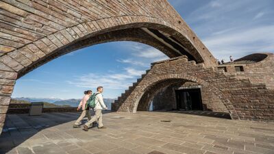 Auf dem Monte Tamaro in Rivera (6802, Kanton Tessin) spazieren Besucher unter den markanten Steinbögen der Kirche Santa Maria degli Angeli mit Blick auf die Alpen.