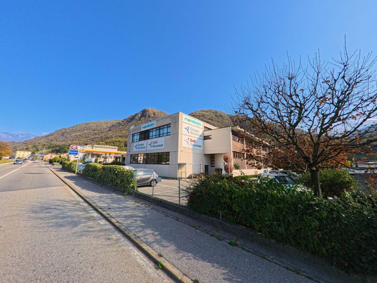 Entlang einer Hauptstrasse steht ein Gewerbegebäude mit Parkplätzen in Monteceneri (6809, Kanton Tessin) vor den bewaldeten Hügeln.