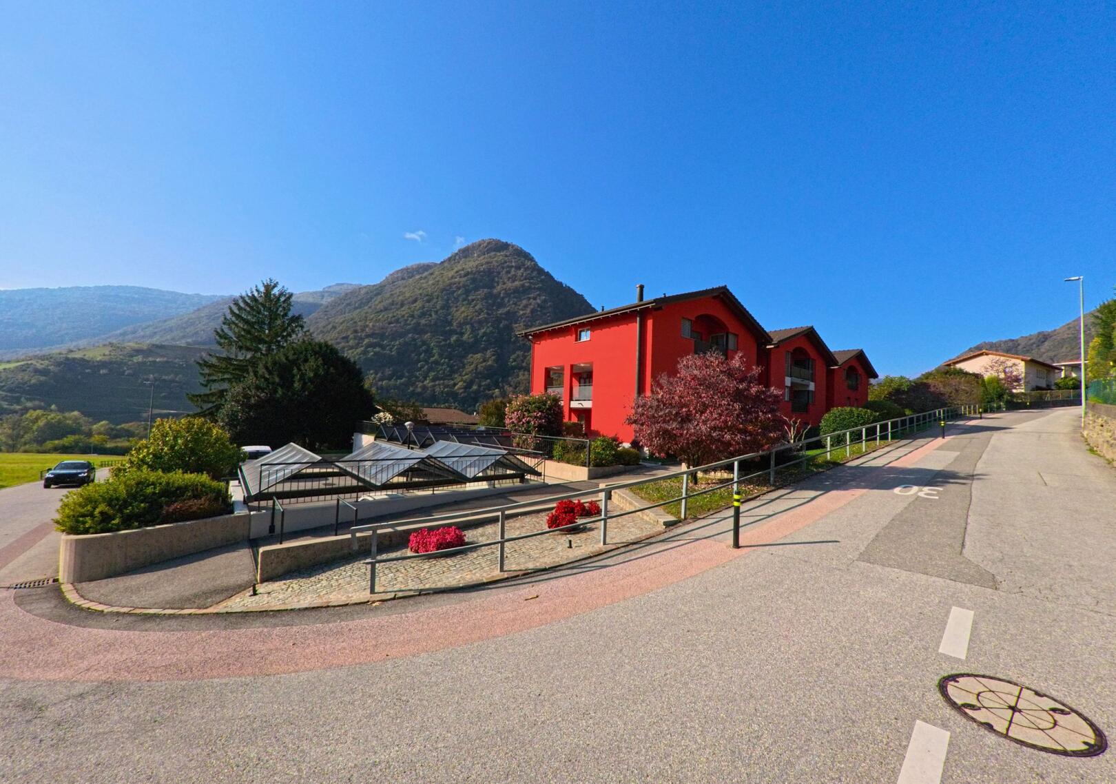 Ein markantes rotes Wohnhaus mit Gartenanlagen liegt an einer leicht ansteigenden Strasse in Monteceneri (6809, Kanton Tessin) vor bewaldeten Berghängen.