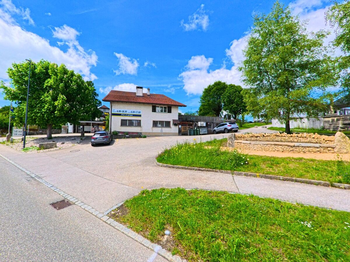 In Montfaucon (2362, Kanton Jura) ist ein Gebäude mit einem Parkplatz und einem Steinbeet an einer Straßenecke zu sehen.