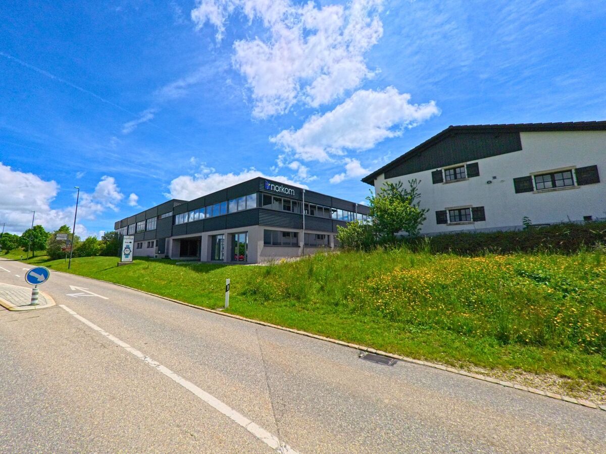In Montfaucon (2362, Kanton Jura) ist ein modernes Gebäude mit einem Firmenlogo an einer Straßenkreuzung sichtbar, daneben ein weiteres Haus und grüne Wiesen.