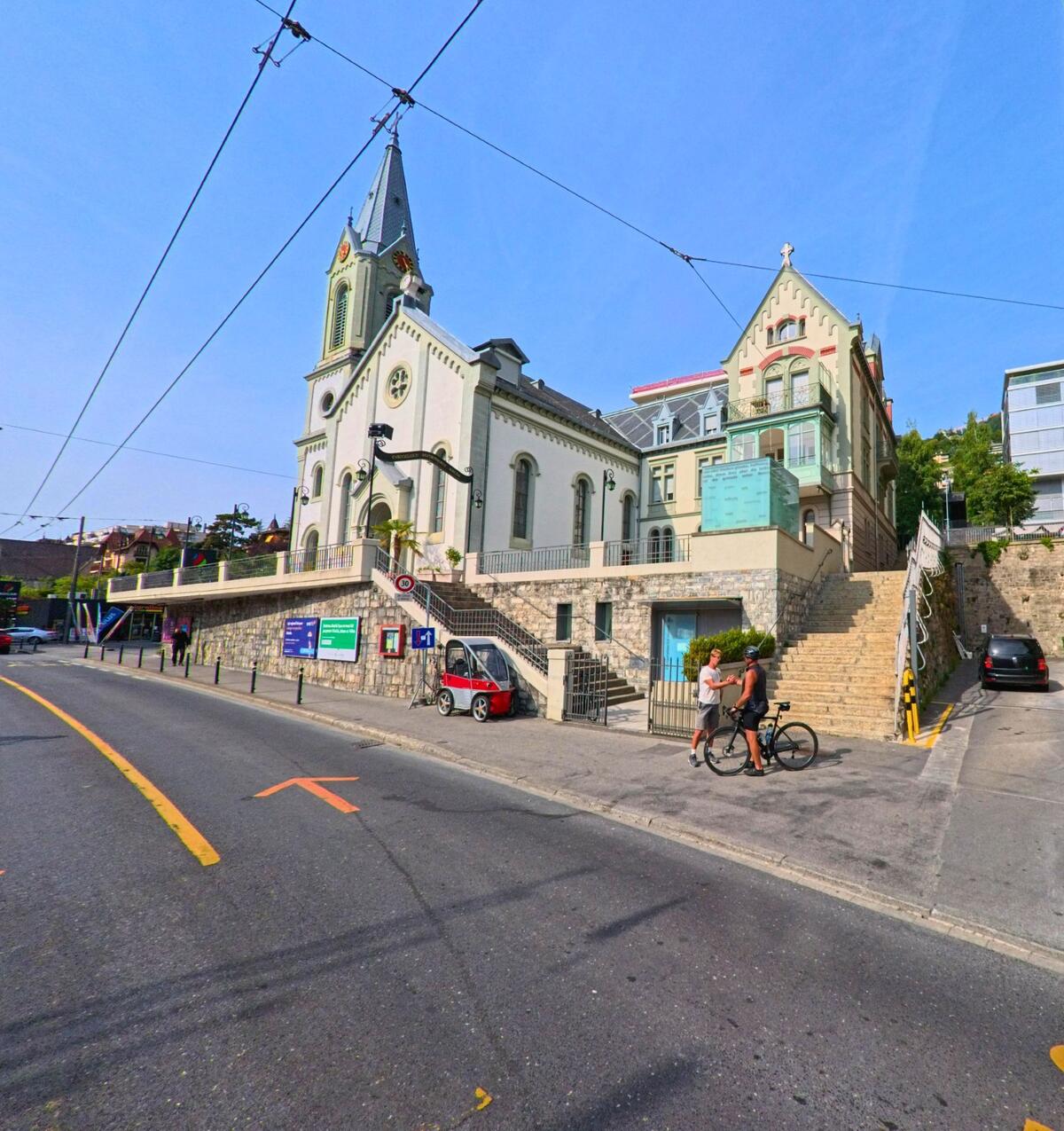Eine Kirche mit hohem Turm und Kreuz steht in Montreux (1820, Kanton Waadt) an einer Strasse, die von Fussgaengern und Radfahrern genutzt wird.