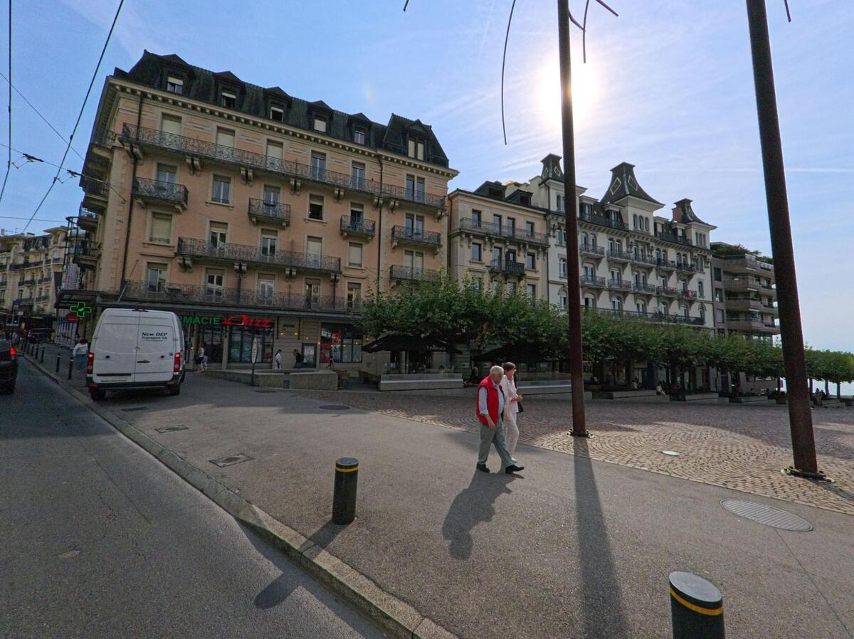 Am Uferplatz in Montreux (1822, Kanton Waadt) stehen historische Wohn- und Hotelgebaeude mit Balkonen, waehrend Passanten bei Sonnenschein vorbeigehen.