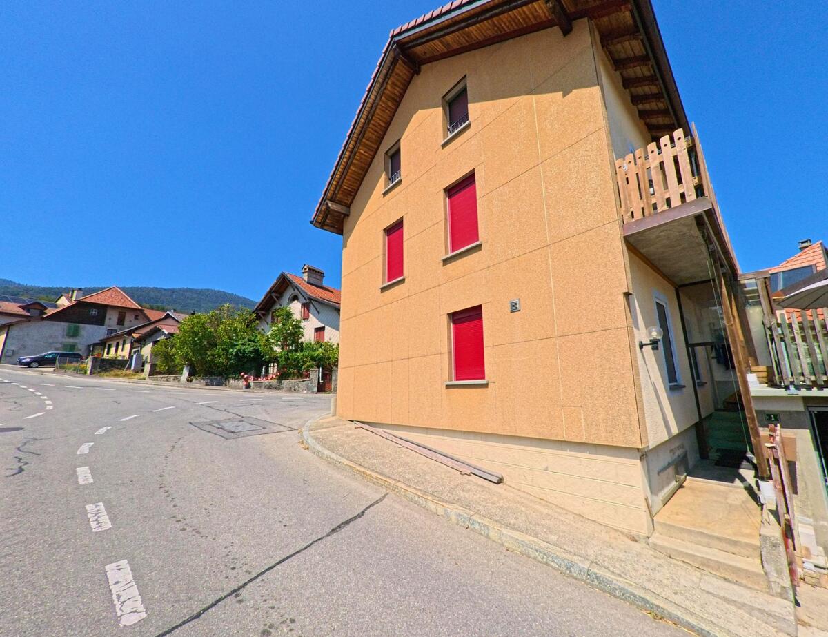 Ein Haus mit roten Fensterlaeden steht an einer Kurve in Montricher (1147, Kanton Waadt), dahinter sind weitere Wohnhaeuser und ein bewaldeter Huegel sichtbar.