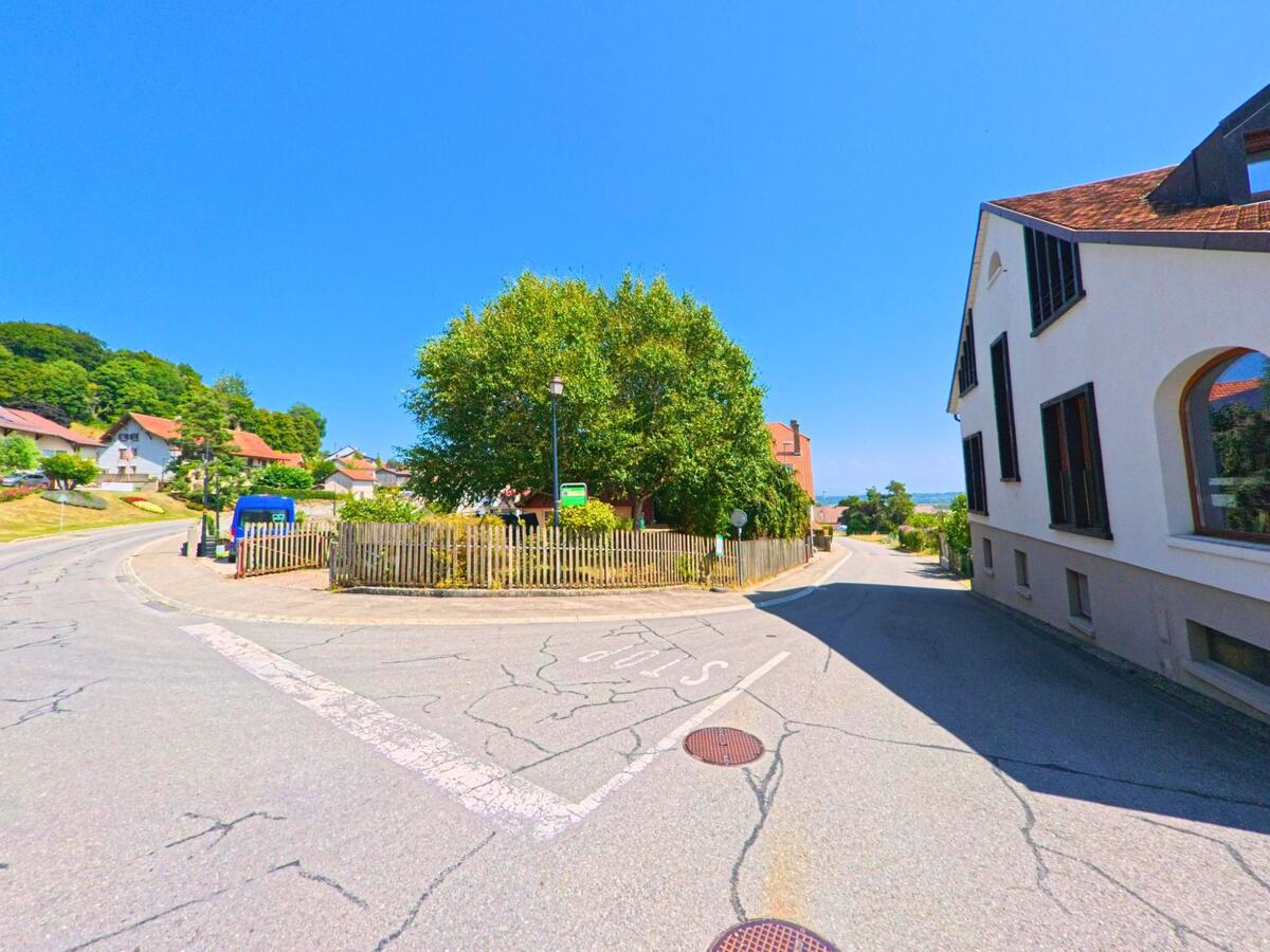 Eine kleine Verkehrsinsel mit Baeumen und Holzzaun praegt eine Kreuzung in Montricher (1147, Kanton Waadt), umgeben von Wohnhaeusern.