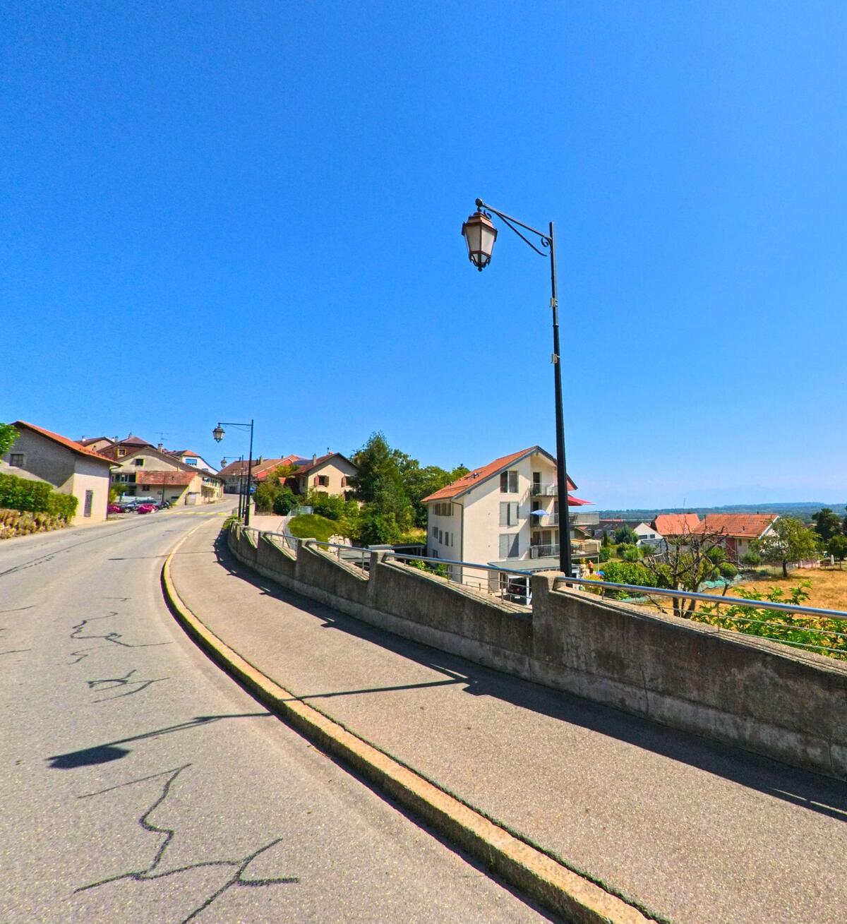 Eine ansteigende Strasse in Montricher (1147, Kanton Waadt) fuehrt an Wohnhaeusern mit roten Daechern vorbei und bietet Fernsicht in die Landschaft.