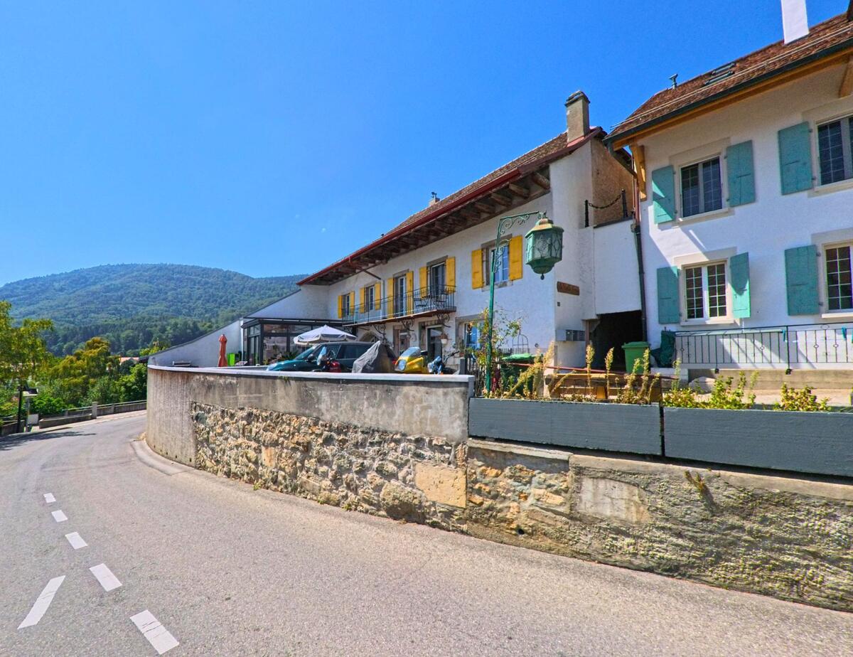 In Montricher (1147, Kanton Waadt) saeumen traditionelle Haeuser mit farbigen Fensterlaeden eine enge Strasse am Hang unterhalb eines bewaldeten Huegels.