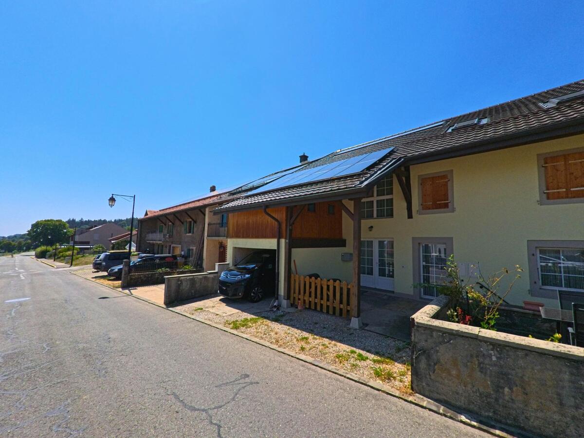 Wohnhaeuser mit Solarpanelen und gedeckten Garagen stehen in Montricher (1147, Kanton Waadt) entlang einer breiten Strasse.