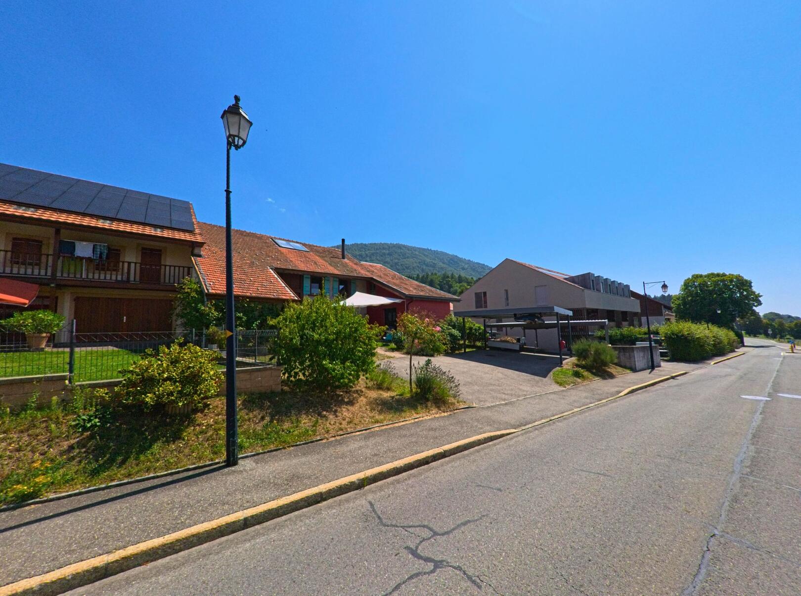 In Montricher (1147, Kanton Waadt) stehen Wohnhaeuser mit roten Daechern und Solarpanelen entlang einer ruhigen Strasse mit Blick auf den bewaldeten Huegel.