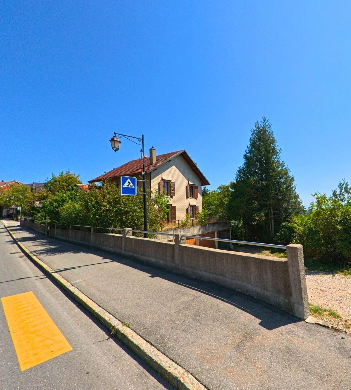In Montricher (1147, Kanton Waadt) steht ein Wohnhaus mit roten Fensterlaeden oberhalb einer Strasse mit markiertem Fussgaengeruebergang.