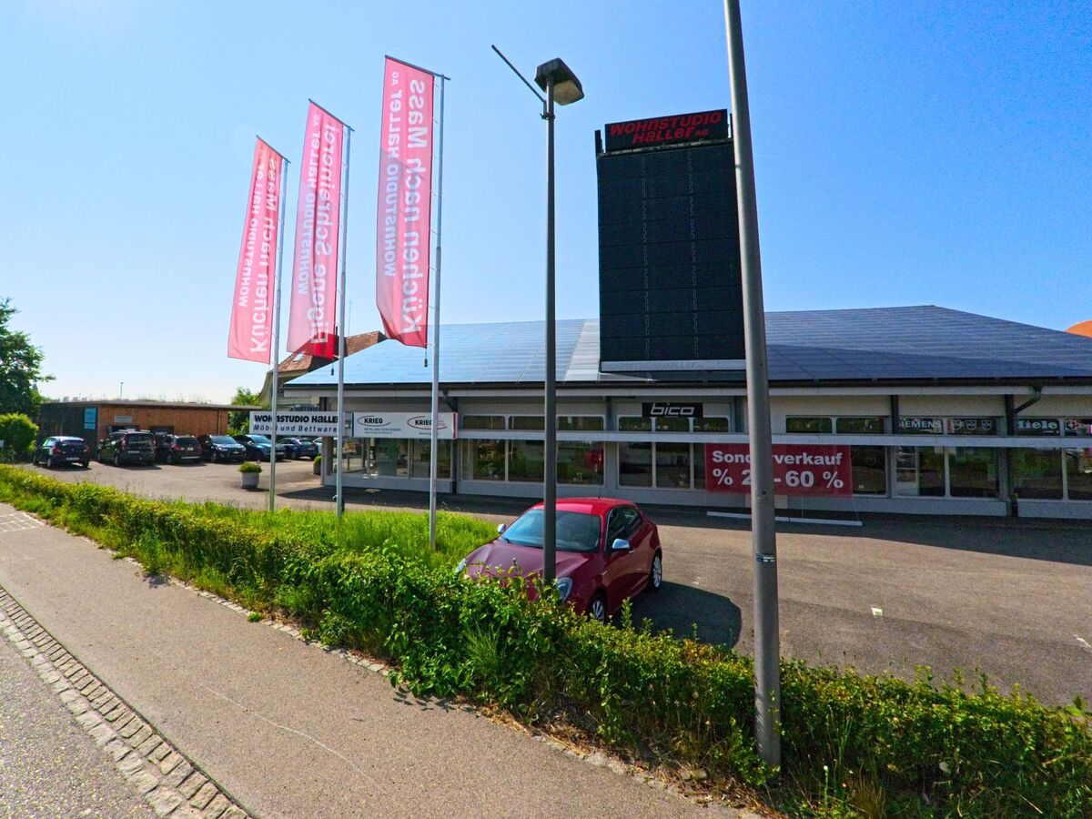 In einem Gewerbegebiet von Moosseedorf (3321, Kanton Bern) steht ein Möbelhaus mit grossen Schaufenstern und Werbefahnen entlang der Strasse im Sonnenlicht.