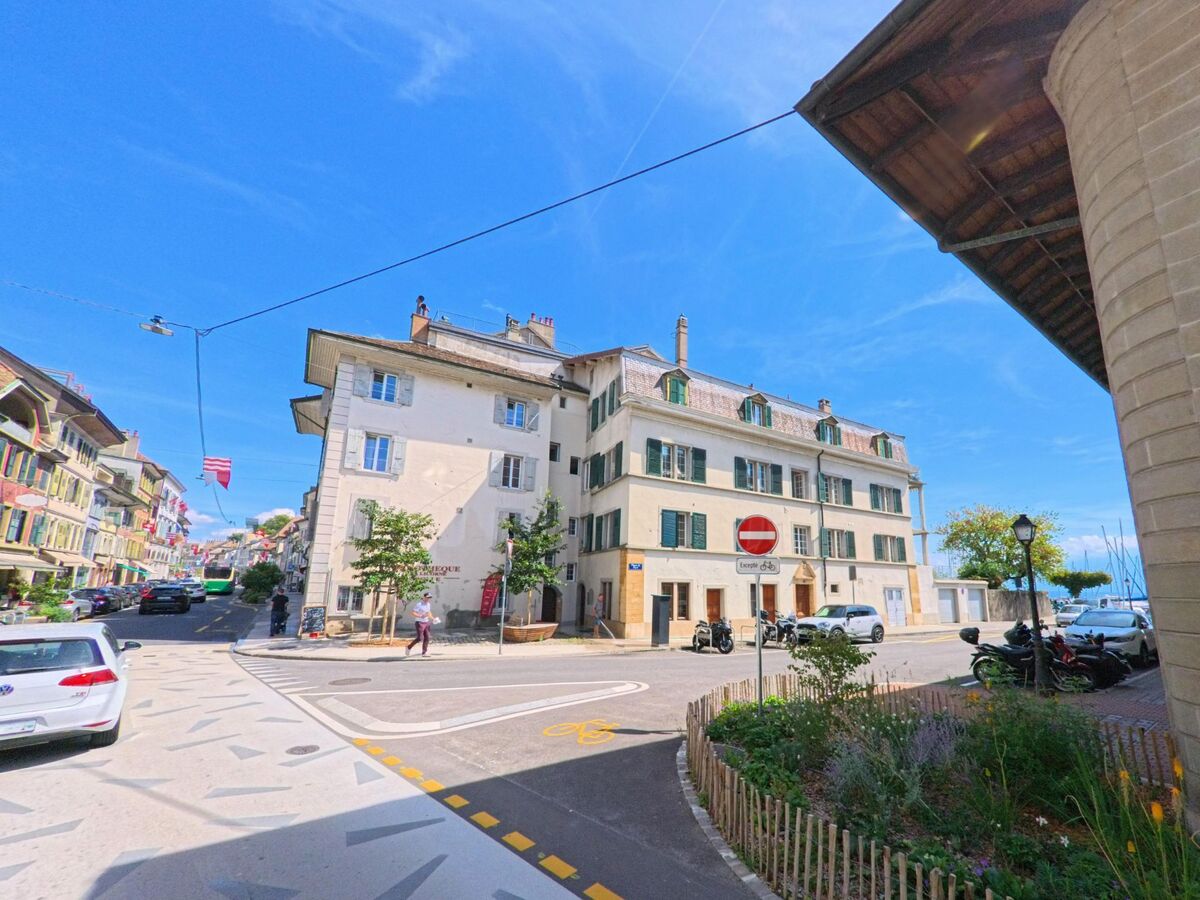 An einer Strassenkreuzung in Morges (1110, Kanton Waadt) stehen helle Wohnhaeuser mit Fensterlaeden, im Hintergrund sind die Masten von Booten am Seeufer sichtbar.