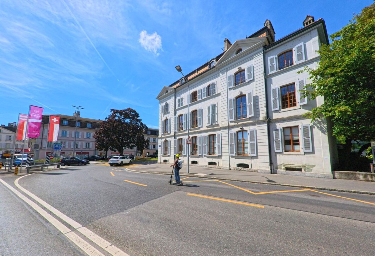 Ein weisses Wohnhaus mit Fensterlaeden steht an einer Kurve in Morges (1110, Kanton Waadt), waehrend ein Mann mit E-Scooter die Strasse ueberquert.