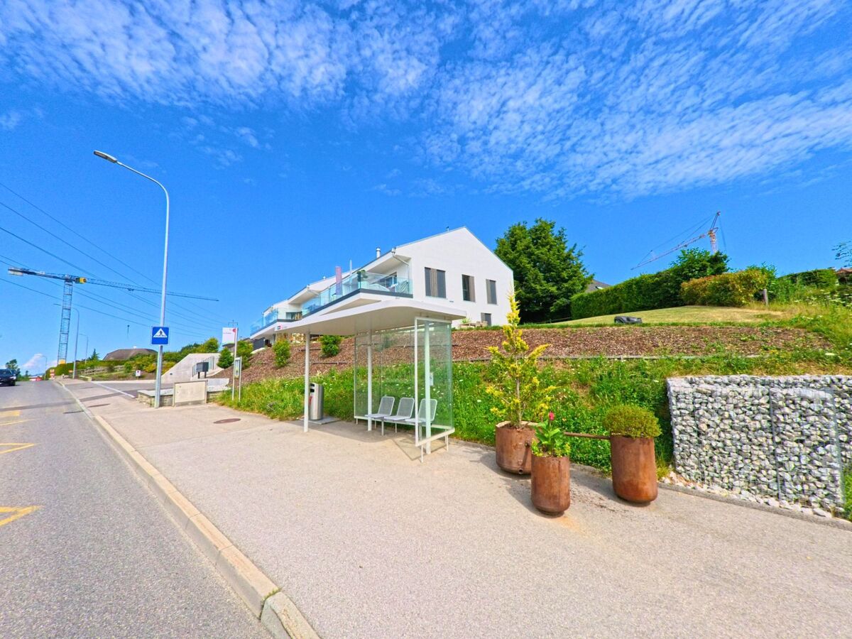 In Morlon (1638, Kanton Freiburg) befindet sich eine moderne Bushaltestelle mit Überdachung am Strassenrand, flankiert von Neubauten und bepflanzten Hängen.