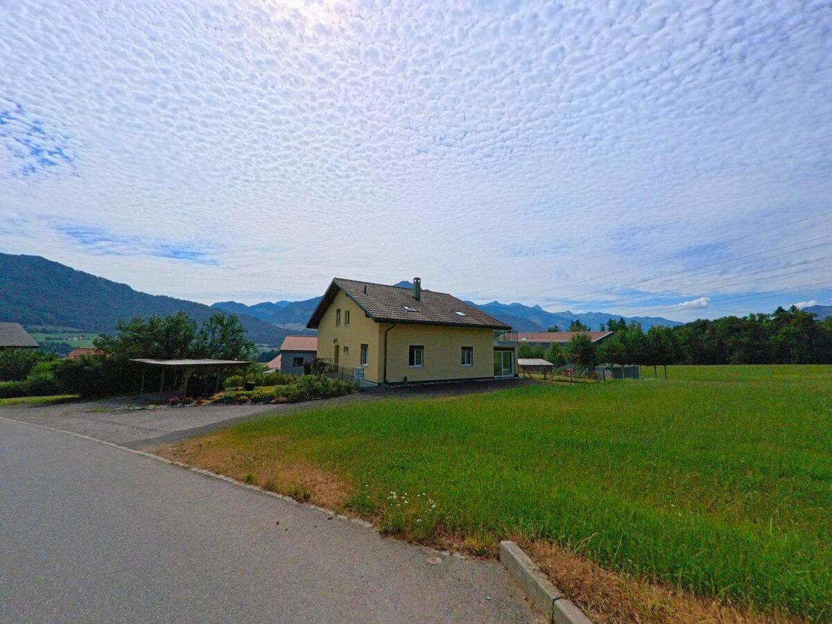 Am Rande von Morlon (1638, Kanton Freiburg) steht ein einzelnes Wohnhaus vor weiter Wiesenlandschaft mit Blick auf die umliegenden Berge.
