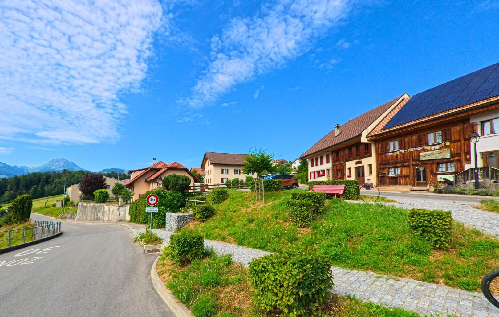 In der Gemeinde Morlon (1638, Kanton Freiburg) verläuft eine ruhige Quartierstrasse an gepflegten Einfamilienhäusern vorbei, während im Vordergrund eine Sitzbank zum Verweilen einlädt.