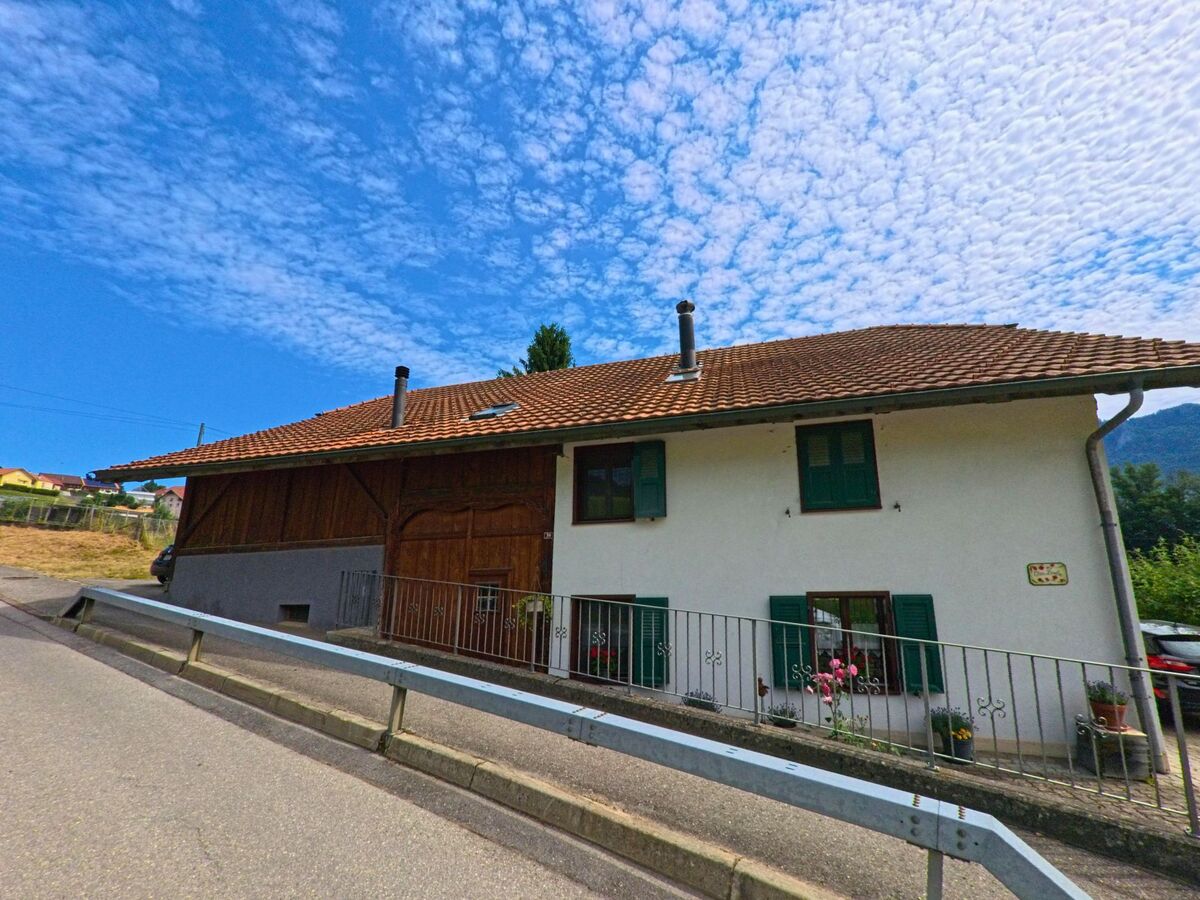 Ein traditionelles Haus mit grünen Fensterläden und gemischter Holz- und Putzfassade steht an einer leicht ansteigenden Strasse in Morlon (1638, Kanton Freiburg).