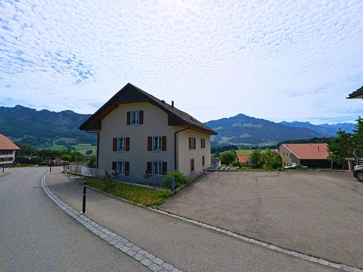 Ein Wohnhaus mit roten Fensterläden steht in Morlon (1638, Kanton Freiburg) am Ende einer Quartierstrasse mit freiem Blick auf das Bergpanorama im Hintergrund.