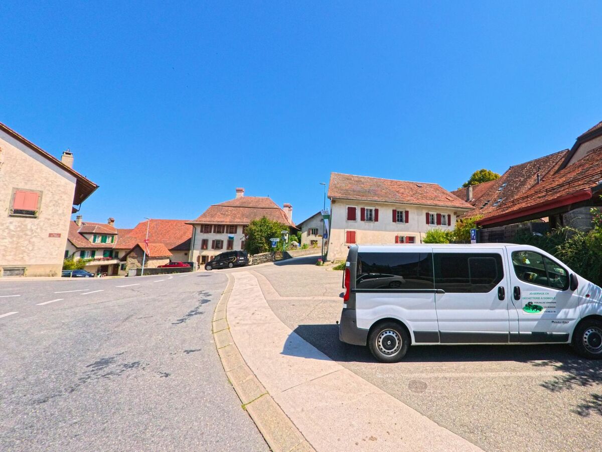 Im Dorfzentrum von Morrens (1054, Kanton Waadt) stehen traditionelle Wohnhaeuser mit roten Fensterlaeden und Ziegeldach entlang einer breiten Strasse.