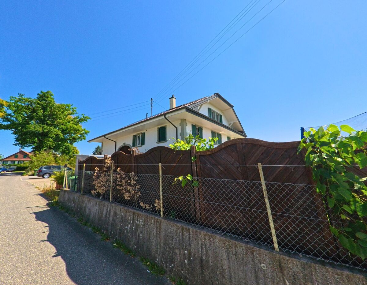 Ein Einfamilienhaus mit gruenen Fensterlaeden steht in Morrens (1054, Kanton Waadt) hinter einem Holzzaun und einem Maschendrahtzaun an einer ruhigen Strasse.