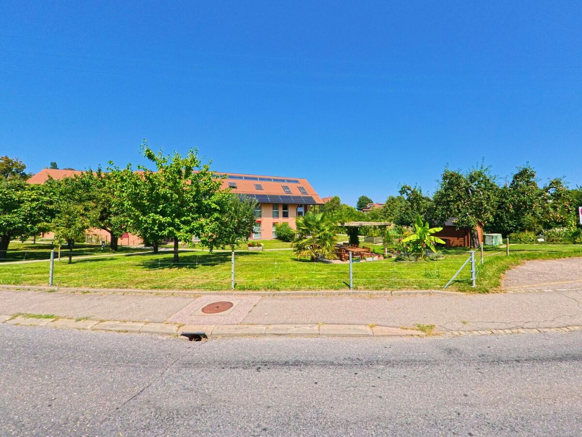 Vor einem grossen Gebaeude mit Solarpaneelen auf dem Dach in Morrens (1054, Kanton Waadt) liegen gepflegte Gruenanlagen mit Baumen und einem kleinen Gartenbereich.