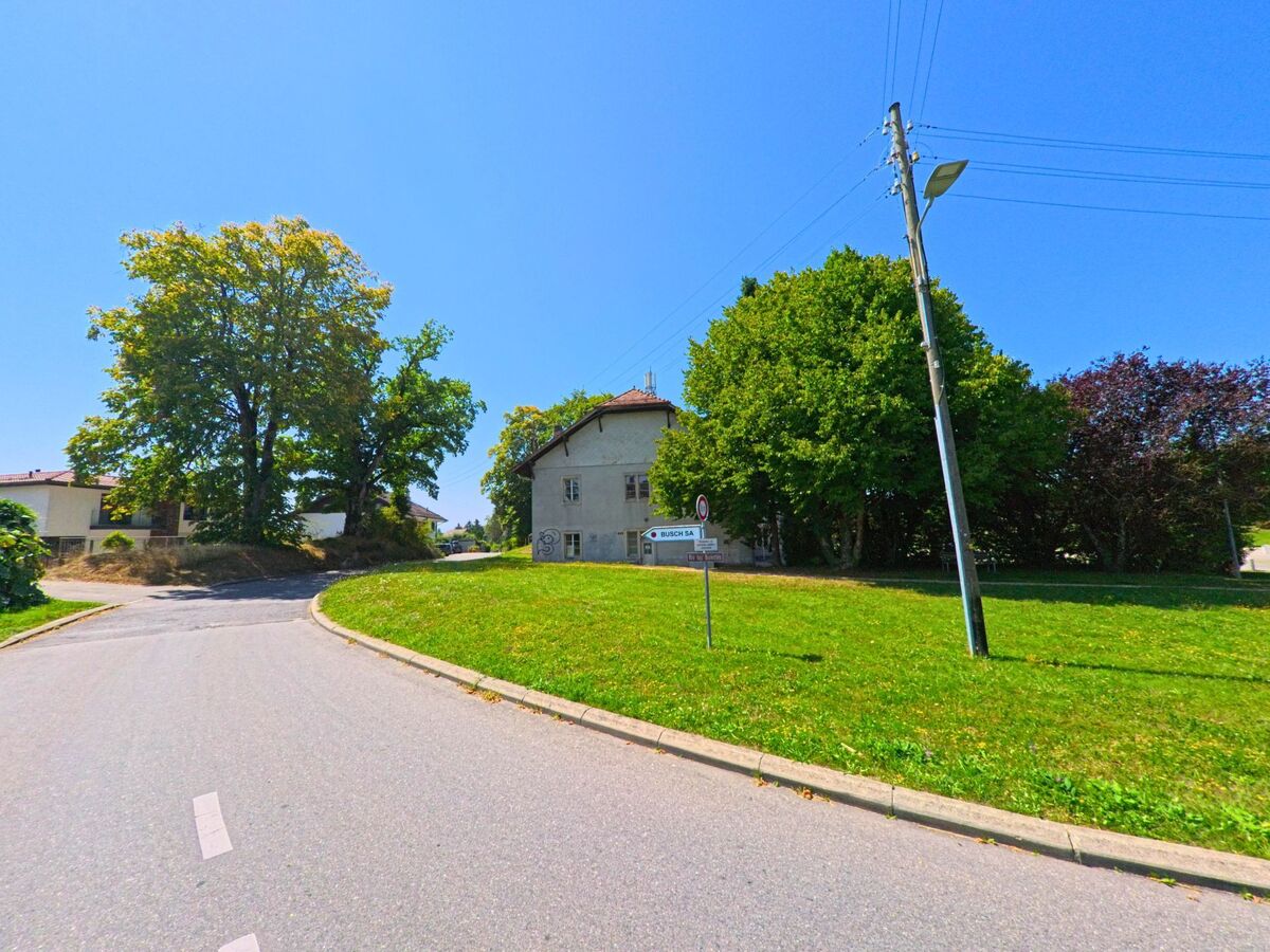An einer kleinen Strassenkreuzung in Morrens (1054, Kanton Waadt) steht ein schlichtes Wohnhaus neben grossen Baeumen und gruener Wiese.