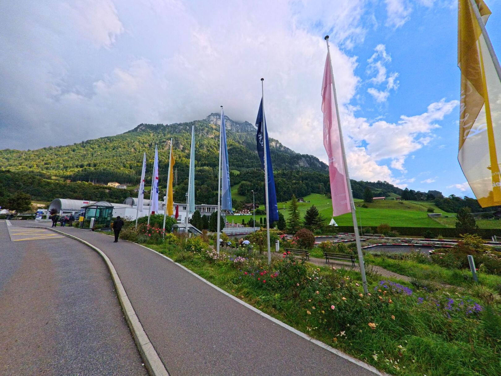 In Morschach (6443, Kanton Schwyz führen farbenfrohe Flaggen entlang einer Straße, die sich vor einer grünen Berglandschaft erstreckt.
