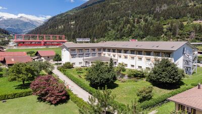 In Quinto (6776, Kanton Tessin) liegt das Motel Gottardo Sud inmitten gruener Wiesen mit Blick auf die umliegenden Berge und die rote Autobahnbruecke im Hintergrund.