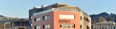 In Wollerau (8832, Kanton Schwyz) steht ein modernes Motelgebäude mit roter Fassade und grosser Terrasse vor der Kulisse bewaldeter Hügel.